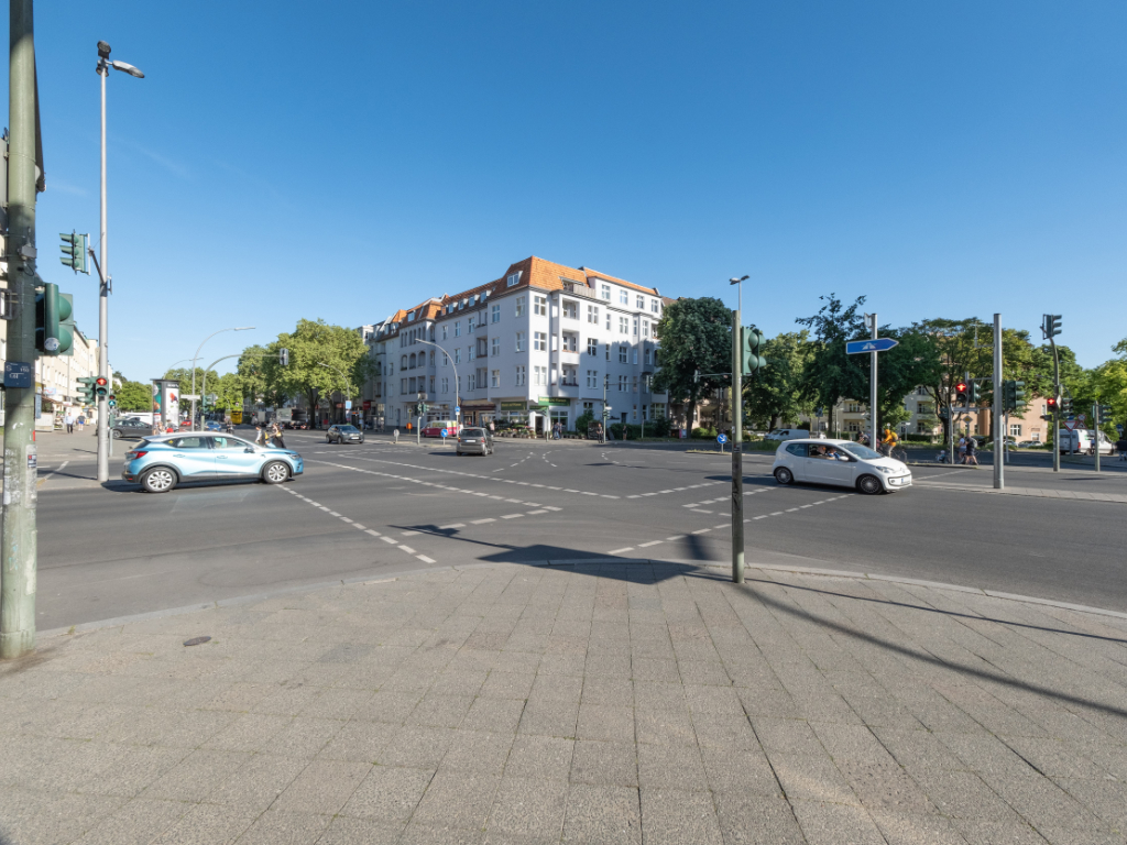 Blick über eine Straßenkreuzung im Sonnenlicht – von einer Straßenecke zur diagonal gegenüberliegenden Ecke. Die Straßen sind mehrspurig mit Autos, Fahrradfahrenden und zu Fuß Gehenden.