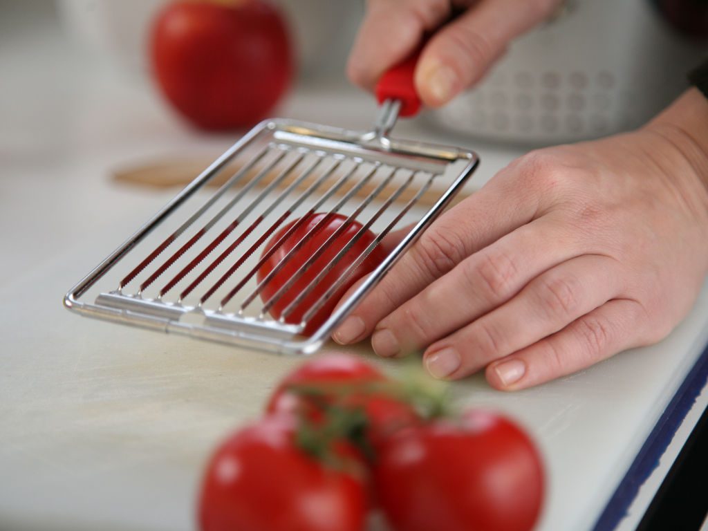 Auf einer weißen Arbeitsfläche wird eine Tomate von einer linken Hand gehalten, während die rechte Hand von oben einen Tomatenschneider ansetzt (ca. handflächengroßer, rechteckiger Metallrahmen, in dem 10 Schneidedrähte gespannt sind, mit rotem Griff).